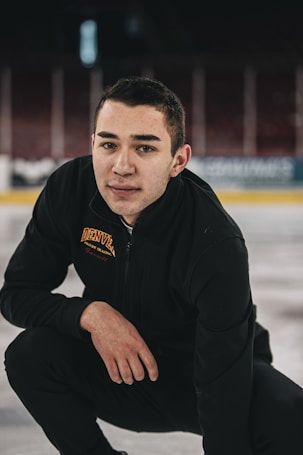 A person kneels on an ice rink wearing black athletic attire with 'Denver' and 'figure skating' embroidered on the jacket. The background shows an empty arena with blurred seats and advertisements.