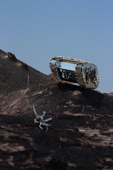 A small, metallic, robotic insect traverses across a dark, textured surface resembling rough bark. In the background, a metallic tank-like structure with intricate details is positioned on the terrain. The sky is clear and blue, providing a stark contrast to the darker elements in the foreground.