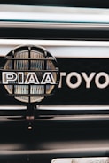 A close-up view of a car's front grill featuring a PIAA auxiliary light with a protective grid and branding, and part of the Toyota emblem visible in the background.