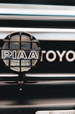 A close-up view of a car's front grill featuring a PIAA auxiliary light with a protective grid and branding, and part of the Toyota emblem visible in the background.