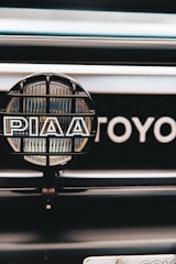 A close-up view of a car's front grill featuring a PIAA auxiliary light with a protective grid and branding, and part of the Toyota emblem visible in the background.
