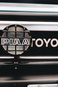 A close-up view of a car's front grill featuring a PIAA auxiliary light with a protective grid and branding, and part of the Toyota emblem visible in the background.