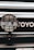 A close-up view of a car's front grill featuring a PIAA auxiliary light with a protective grid and branding, and part of the Toyota emblem visible in the background.