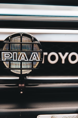 A close-up view of a car's front grill featuring a PIAA auxiliary light with a protective grid and branding, and part of the Toyota emblem visible in the background.