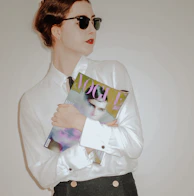 A person with red lipstick wearing sunglasses and a white shirt is holding a fashion magazine against a neutral background. The magazine cover features a portrait and the word 'VOGUE' in large letters.
