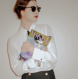 A person with red lipstick wearing sunglasses and a white shirt is holding a fashion magazine against a neutral background. The magazine cover features a portrait and the word 'VOGUE' in large letters.