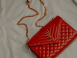 A red quilted leather handbag with a gold chain strap and metallic logo accent rests on a light-colored fabric surface.