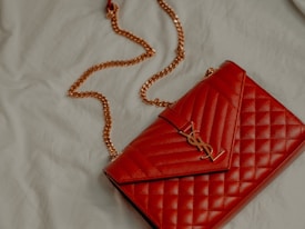 A red quilted leather handbag with a gold chain strap and metallic logo accent rests on a light-colored fabric surface.