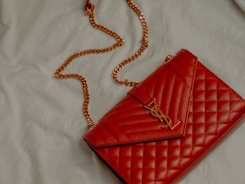 A red quilted leather handbag with a gold chain strap and metallic logo accent rests on a light-colored fabric surface.