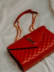 A luxurious red leather handbag with a quilted pattern and a prominent gold logo. The bag features a gold chain strap and is placed on a soft, light-colored fabric background.