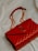 A luxurious red leather handbag with a quilted pattern and a prominent gold logo. The bag features a gold chain strap and is placed on a soft, light-colored fabric background.