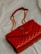A luxurious red leather handbag with a quilted pattern and a prominent gold logo. The bag features a gold chain strap and is placed on a soft, light-colored fabric background.