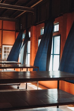 Tables are arranged in rows inside a room with orange walls and large windows draped with dark blue curtains. Sunlight streams through the windows, creating shadows on the tiled floor and tables.