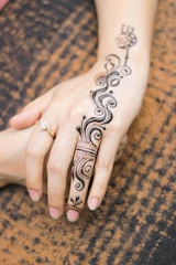 A close-up view of a hand adorned with intricate henna designs that feature swirling patterns and floral motifs extending from the wrist to the finger. The skin appears light and is complemented by a delicate ring on one finger, set against a textured brown fabric background.