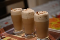 Three tall glasses filled with a creamy, frothy beverage are neatly arranged on a tray. The drinks have a light brown color topped with foam and are garnished with almond slices.