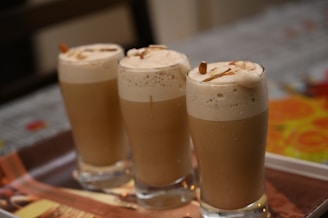 Three tall glasses filled with a creamy, frothy beverage are neatly arranged on a tray. The drinks have a light brown color topped with foam and are garnished with almond slices.
