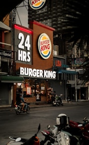 A Burger King restaurant operates 24 hours and is situated in an urban setting. The establishment displays illuminated signage with the brand's logo. Several motorbikes are visible parked in front, and a person rides past on a motorbike on the street. Additional small stores and signage are seen nearby.