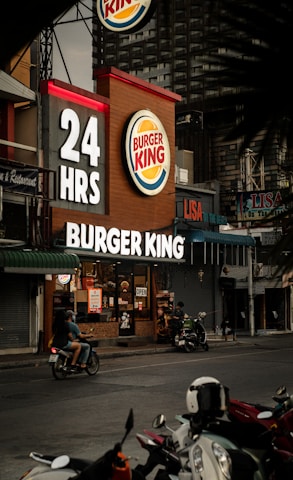 A Burger King restaurant operates 24 hours and is situated in an urban setting. The establishment displays illuminated signage with the brand's logo. Several motorbikes are visible parked in front, and a person rides past on a motorbike on the street. Additional small stores and signage are seen nearby.