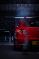 A red car with visible exhaust fumes is parked in a dimly lit parking garage. The back of the car is prominently displayed with illuminated taillights and markings in Japanese on the rear windshield.