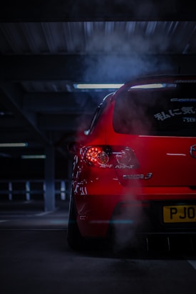 A red car with visible exhaust fumes is parked in a dimly lit parking garage. The back of the car is prominently displayed with illuminated taillights and markings in Japanese on the rear windshield.