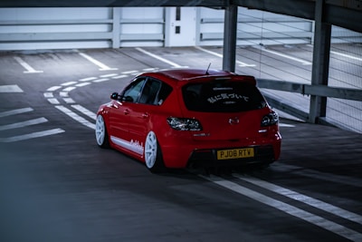 A red car is parked on the top level of a parking garage. Its design features a sporty look with prominent white wheels and decals on the side. The rear window has several stickers, including text in Japanese. The setting appears to be dimly lit, emphasizing the car's vibrant color against the subdued background.