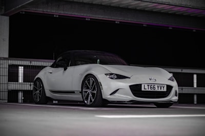 A sleek white sports car is parked indoors under an overhead structure. The car features a low stance and stylish design with visible 'Recaro' branding on the side. It is captured against a dark background, highlighting its elegant contours and glossy finish.
