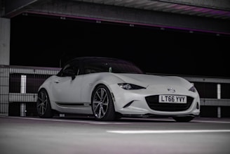A sleek white sports car is parked indoors under an overhead structure. The car features a low stance and stylish design with visible 'Recaro' branding on the side. It is captured against a dark background, highlighting its elegant contours and glossy finish.