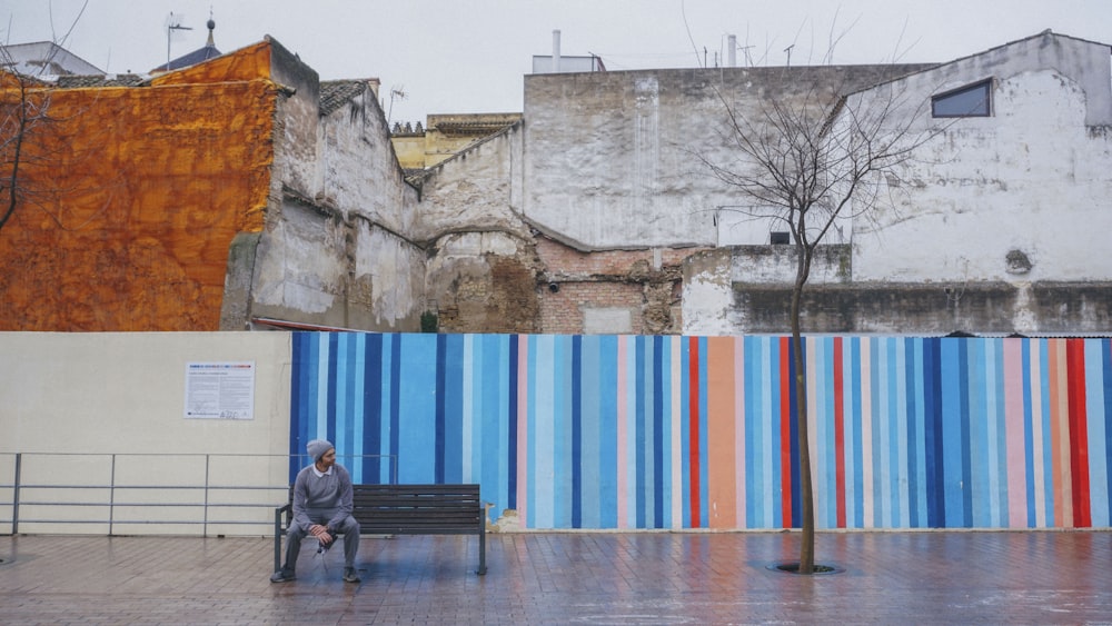 man in black jacket sitting on bench near blue and white wall during daytime