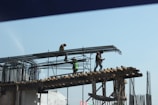 Construction workers are standing on a scaffold, installing steel reinforcement bars on a building under construction. The workers are equipped with safety gear, including helmets and harnesses, while carefully maneuvering the bars into place.