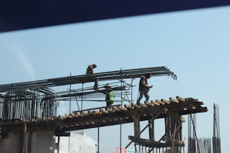 Construction workers are standing on a scaffold, installing steel reinforcement bars on a building under construction. The workers are equipped with safety gear, including helmets and harnesses, while carefully maneuvering the bars into place.