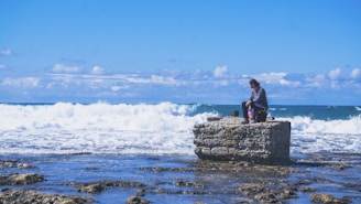 A person is sitting on a rocky platform by the sea, surrounded by gentle waves and an expansive blue sky with scattered clouds. The scene conveys a sense of tranquility and contemplation.