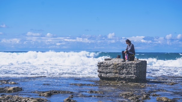 A person is sitting on a rocky platform by the sea, surrounded by gentle waves and an expansive blue sky with scattered clouds. The scene conveys a sense of tranquility and contemplation.