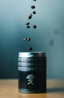 Several coffee beans are captured in mid-air, falling into a decorative black canister with intricate patterns and Asian script. The background is softly blurred, putting focus on the canister and the beans.