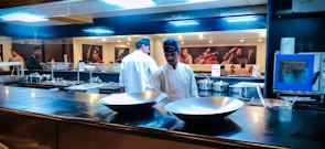 A commercial kitchen setting with chefs in white uniforms and black hats working at the counter. The kitchen is equipped with shiny metal bowls, cooking pots, and utensils. The background features a wall adorned with vibrant photographs of musicians.