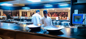A commercial kitchen setting with chefs in white uniforms and black hats working at the counter. The kitchen is equipped with shiny metal bowls, cooking pots, and utensils. The background features a wall adorned with vibrant photographs of musicians.