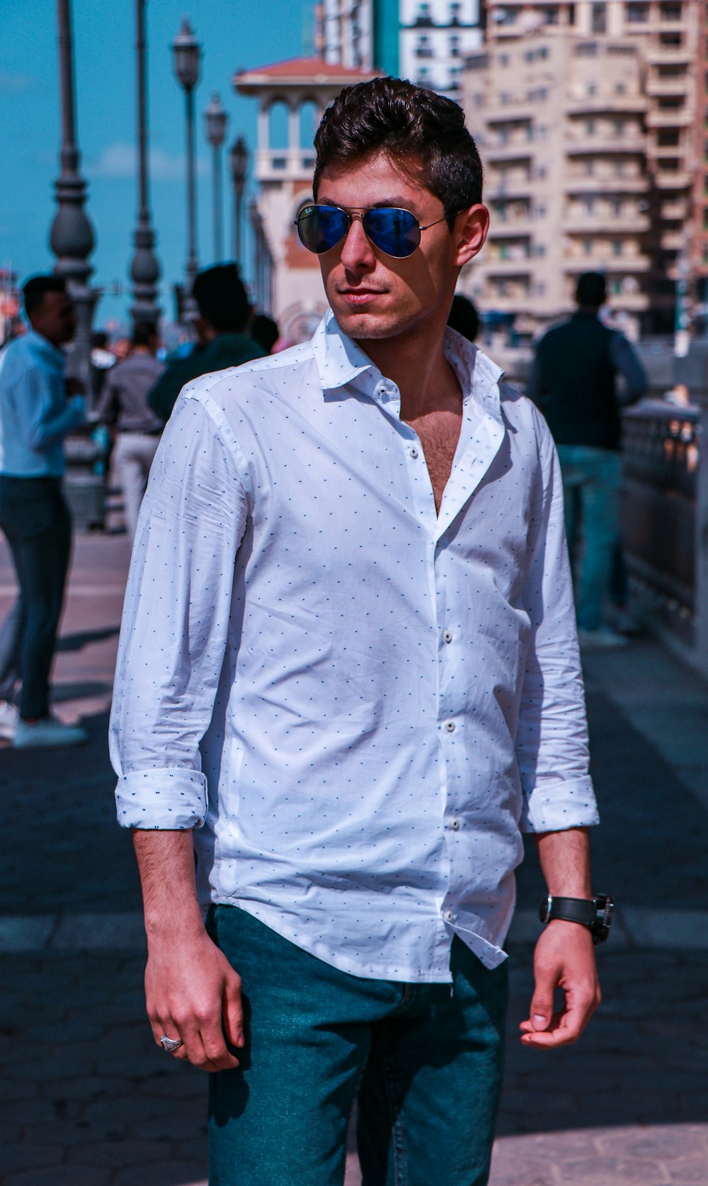青いドレスシャツと青いデニムジーンズを着たサングラスをかけた男性が昼間、歩道に立っている