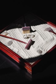 A small zen garden with carefully raked white sand, featuring a few smooth stones and small wooden tools including a rake and a brush. The scene is lit by a soft light creating shadows, and the garden is set in a square wooden frame.
