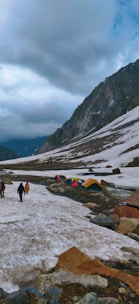 A snow-covered landscape with a mountain range in the background. Colorful tents are pitched along a narrow stream, and a few people are walking along the snowy path. The sky is overcast, adding a moody atmosphere to the scene.