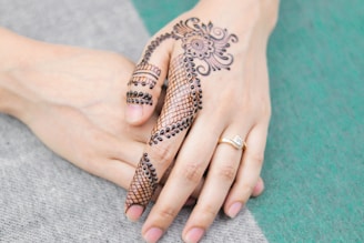 A hand adorned with intricate henna designs, featuring floral and paisley patterns, held by another hand. The henna covers the fingers and back of the hand in a detailed and artistic manner. A gold ring with a small gemstone is worn on the ring finger.