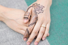 A hand adorned with intricate henna designs, featuring floral and paisley patterns, held by another hand. The henna covers the fingers and back of the hand in a detailed and artistic manner. A gold ring with a small gemstone is worn on the ring finger.