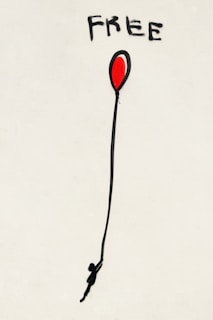 A piece of street art featuring the word 'FREE' painted in bold black letters above a red balloon. The balloon has a long black string with a silhouette of a person hanging onto it, against a plain white backdrop.