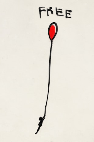 A piece of street art featuring the word 'FREE' painted in bold black letters above a red balloon. The balloon has a long black string with a silhouette of a person hanging onto it, against a plain white backdrop.