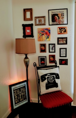 A corner of a room featuring a variety of framed artworks on the wall, including colorful abstract paintings and a magazine cover. There is a tall, vintage-style lamp next to a metal chair with a cushion displaying an old rotary telephone design. On the floor, a framed artwork is leaning against the wall. The chair cushion is red, adding a pop of color.