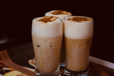 Three glasses of iced coffee topped with a thick layer of frothy cream and cinnamon sprinkled on top. The beverages are served in tall, clear glasses set against a dark background, creating a cozy and inviting atmosphere.