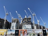 Several large construction cranes are positioned around high-rise buildings under construction. The structures are at different stages of completion, surrounded by various scaffolding and construction materials. Advertising banners at the foreground promote upcoming developments in the area.