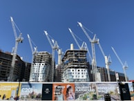 Several large construction cranes are positioned around high-rise buildings under construction. The structures are at different stages of completion, surrounded by various scaffolding and construction materials. Advertising banners at the foreground promote upcoming developments in the area.