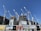 Several large construction cranes are positioned around high-rise buildings under construction. The structures are at different stages of completion, surrounded by various scaffolding and construction materials. Advertising banners at the foreground promote upcoming developments in the area.