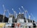 Several large construction cranes are positioned around high-rise buildings under construction. The structures are at different stages of completion, surrounded by various scaffolding and construction materials. Advertising banners at the foreground promote upcoming developments in the area.