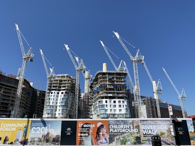 Several large construction cranes are positioned around high-rise buildings under construction. The structures are at different stages of completion, surrounded by various scaffolding and construction materials. Advertising banners at the foreground promote upcoming developments in the area.