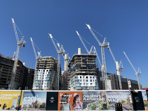 Several large construction cranes are positioned around high-rise buildings under construction. The structures are at different stages of completion, surrounded by various scaffolding and construction materials. Advertising banners at the foreground promote upcoming developments in the area.